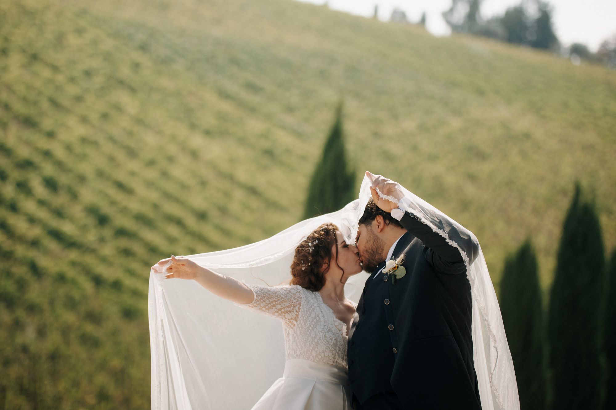 A bride and groom kissing beneath a lifted veil on a green hillside