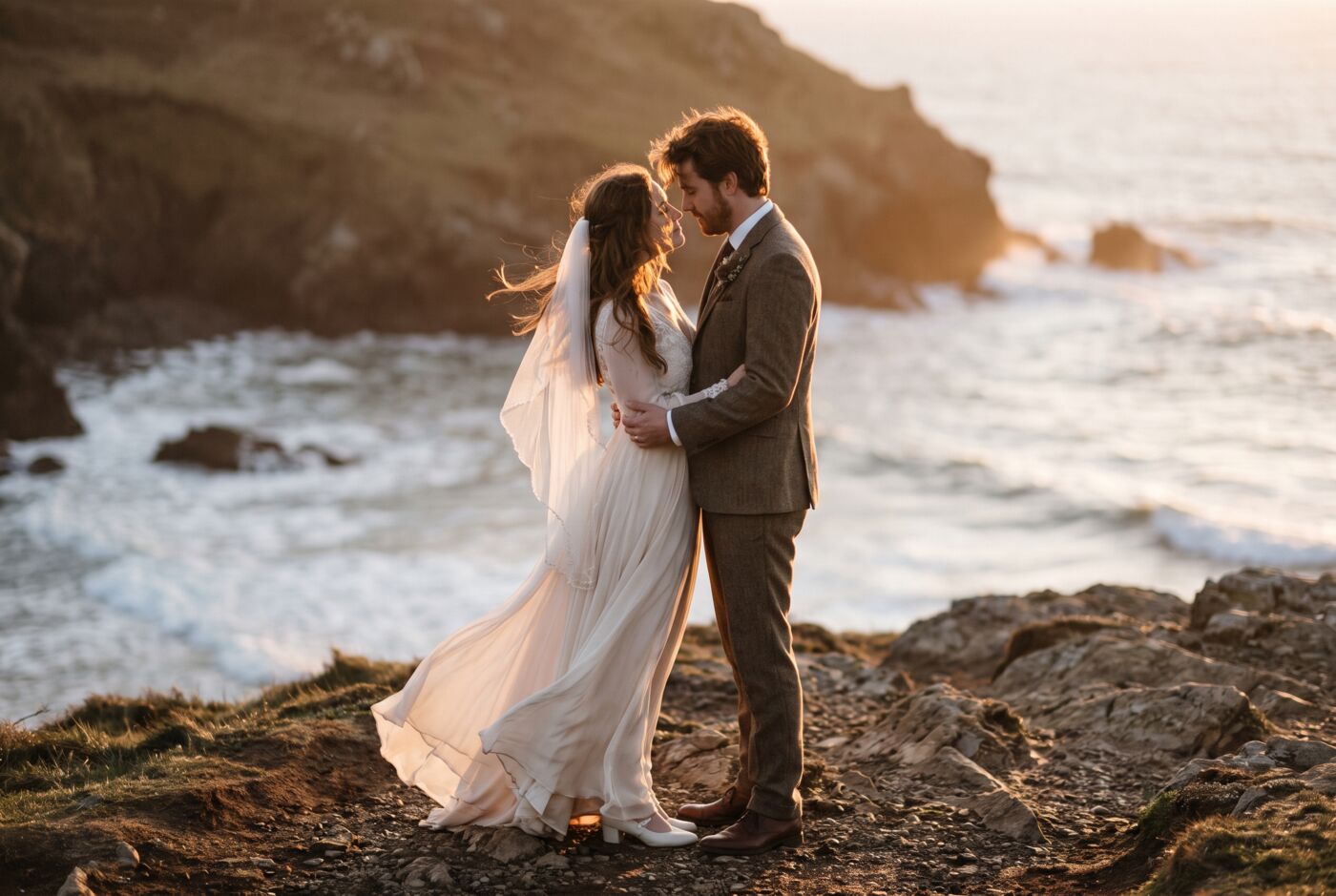 A bride and groom standing close on a windswept coastal cliff at dusk